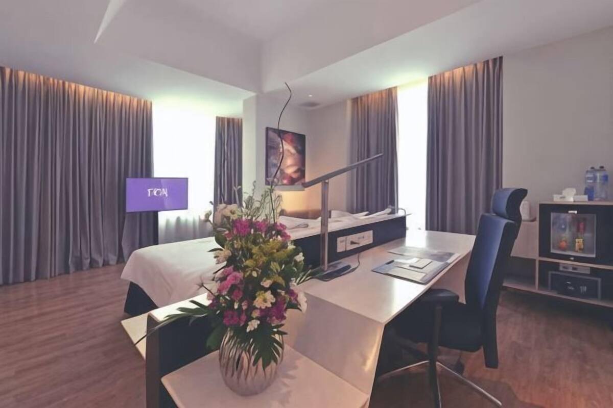 Ett hotellrum med en säng, ett skrivbord, en stol, en tv och en dekorativ vas.