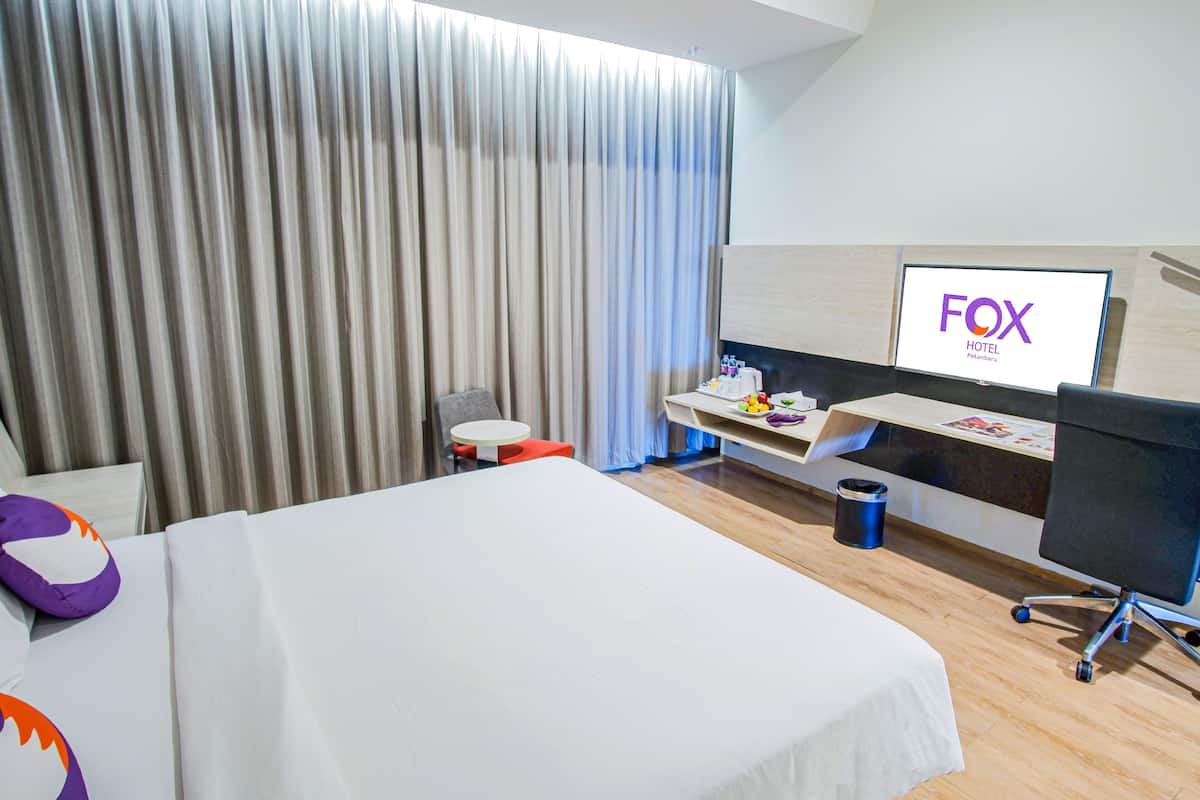Ett hotellrum med en säng, ett skrivbord med en dator, en tv som visar hotellets namn och en gardin.