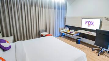 Una habitación de hotel con una cama, un escritorio con computadora, un televisor mostrando el nombre del hotel y una cortina.
