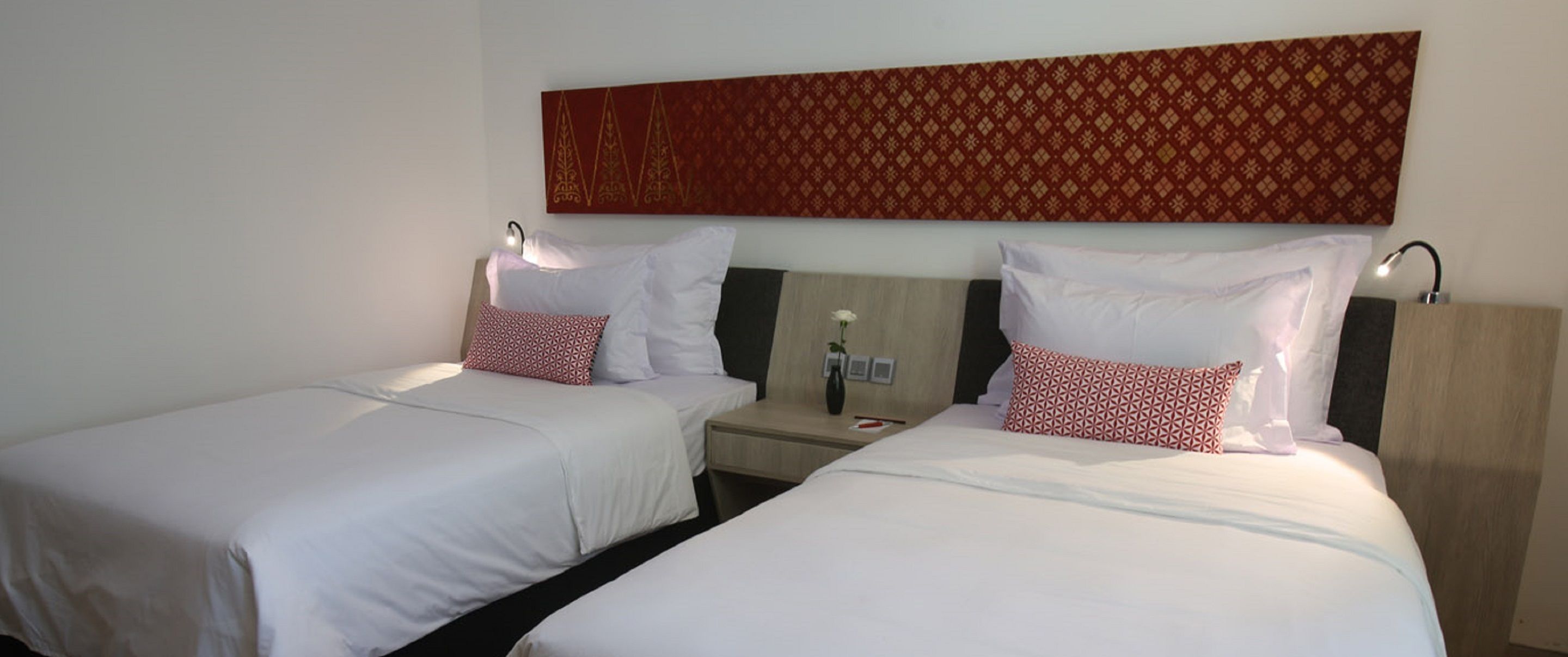 Habitación de hotel con dos camas individuales, cada una con ropa blanca y almohadas estampadas.  Entre las camas hay una mesita de noche con un jarrón y una lámpara.