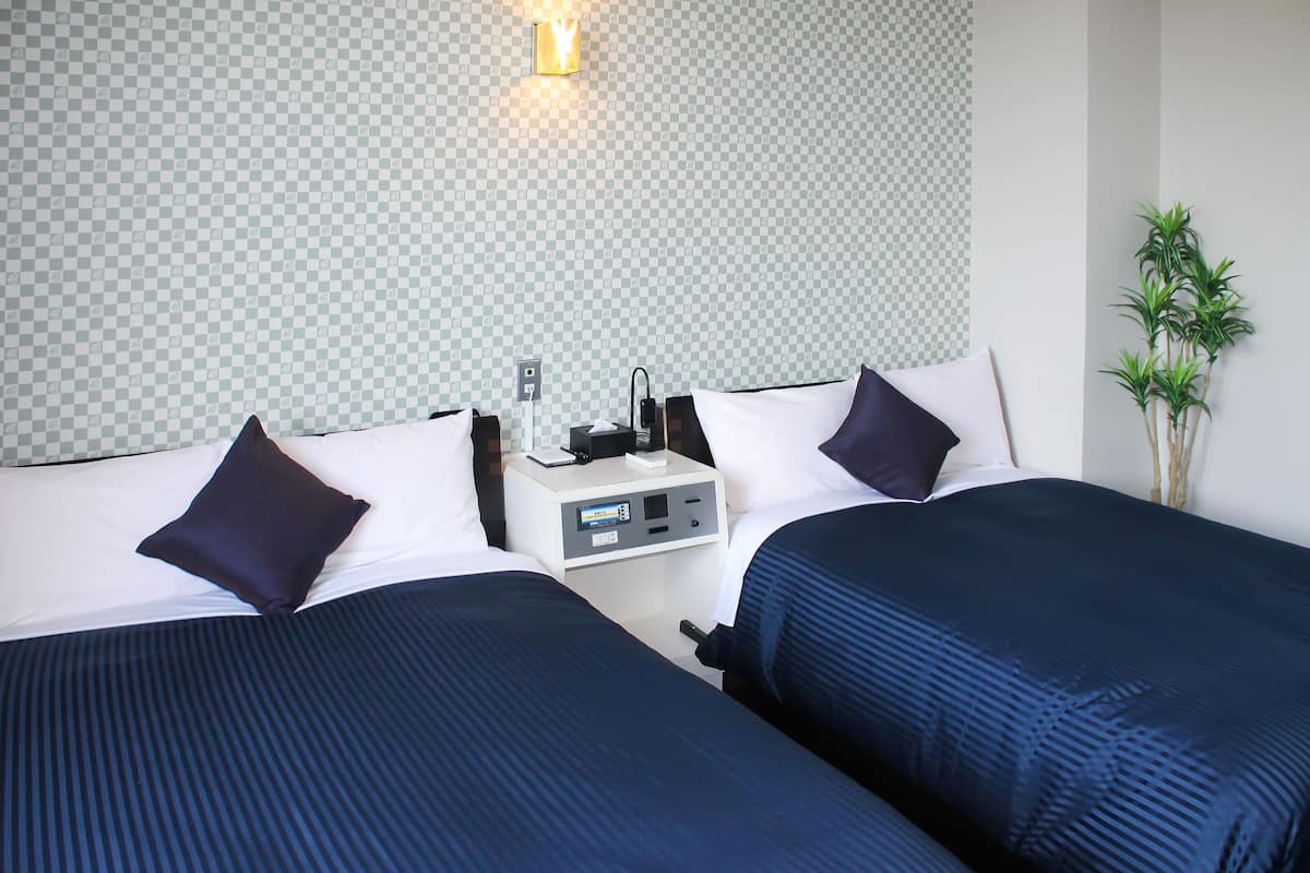 Habitación de hotel con dos camas, una mesita de noche, una lámpara y una planta.