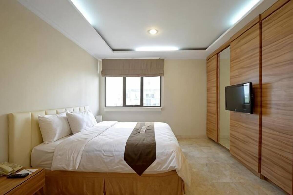Een slaapkamer met een bed, een aan de muur bevestigde televisie, een raam met gordijnen en een houten garderobe.