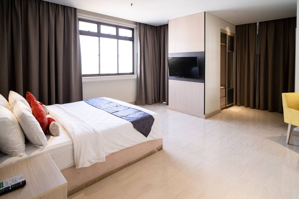 A modern hotel room with a large bed, a TV, a wardrobe, and a seating area.