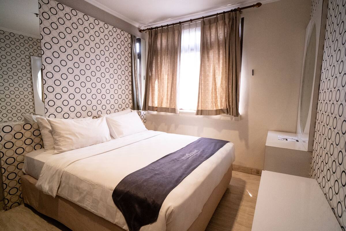 A hotel room with a large bed, a bedside table, a window with curtains, and patterned wallpaper.