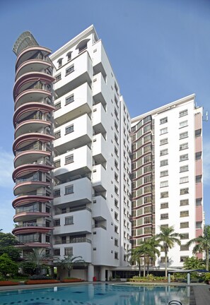 Een meerlaags hotel met een opvallend gebogen balkon aan de linkerkant, een zwembadgedeelte met palmbomen en een helderblauwe lucht.