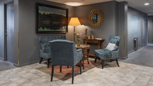 A hotel lobby with a seating area featuring two upholstered chairs, a round wooden table, a lamp, and a framed painting on the wall. - Hotell Leksand (Leksand)