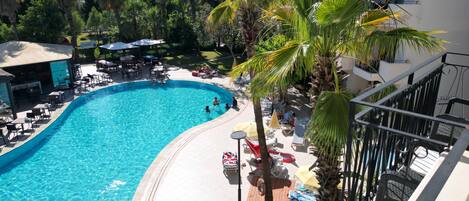 A swimming pool with lounge chairs and umbrellas, surrounded by palm trees and a building with balconies.