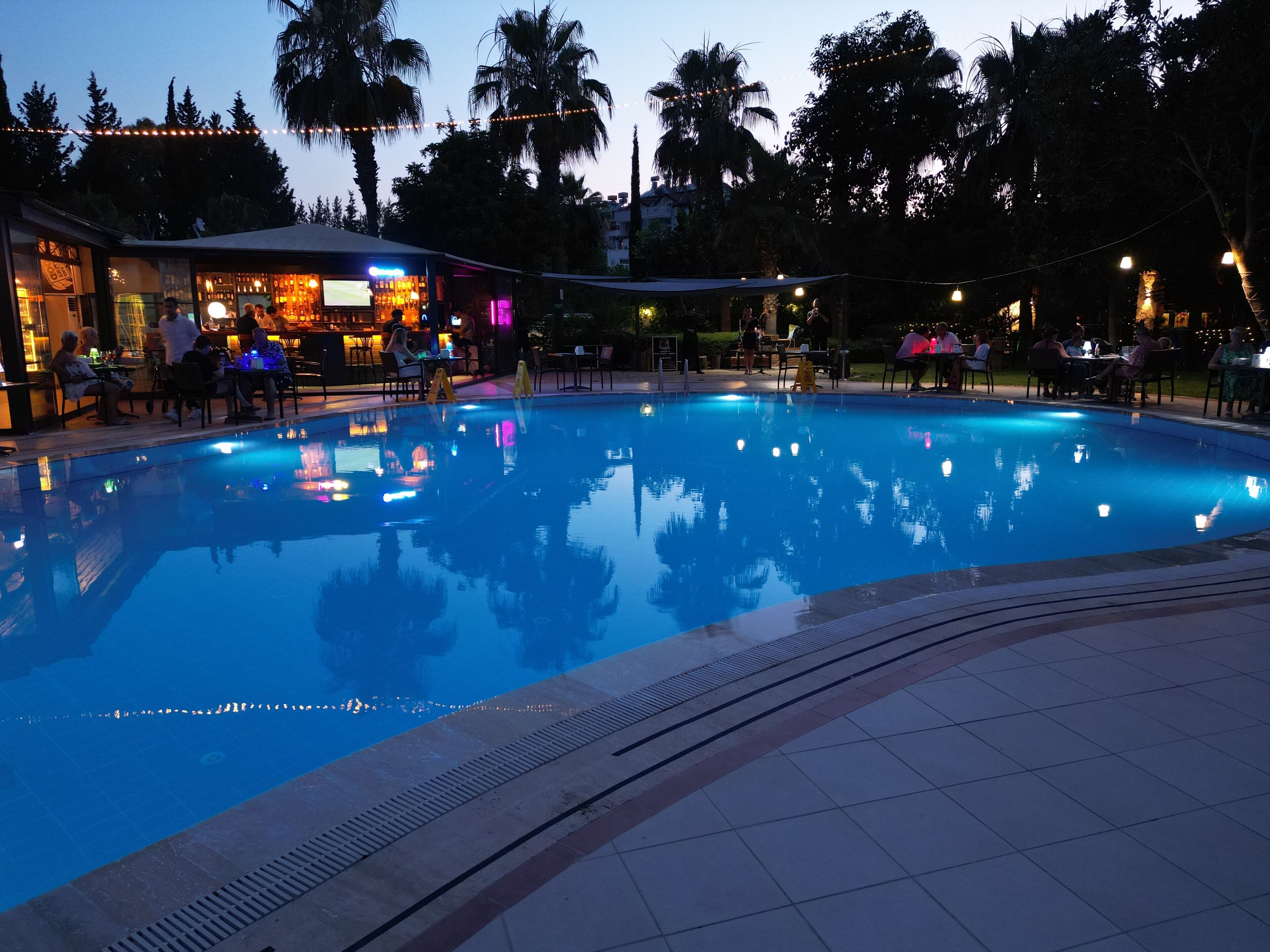 Piscina con bar y zona de estar, rodeada de palmeras y edificios.