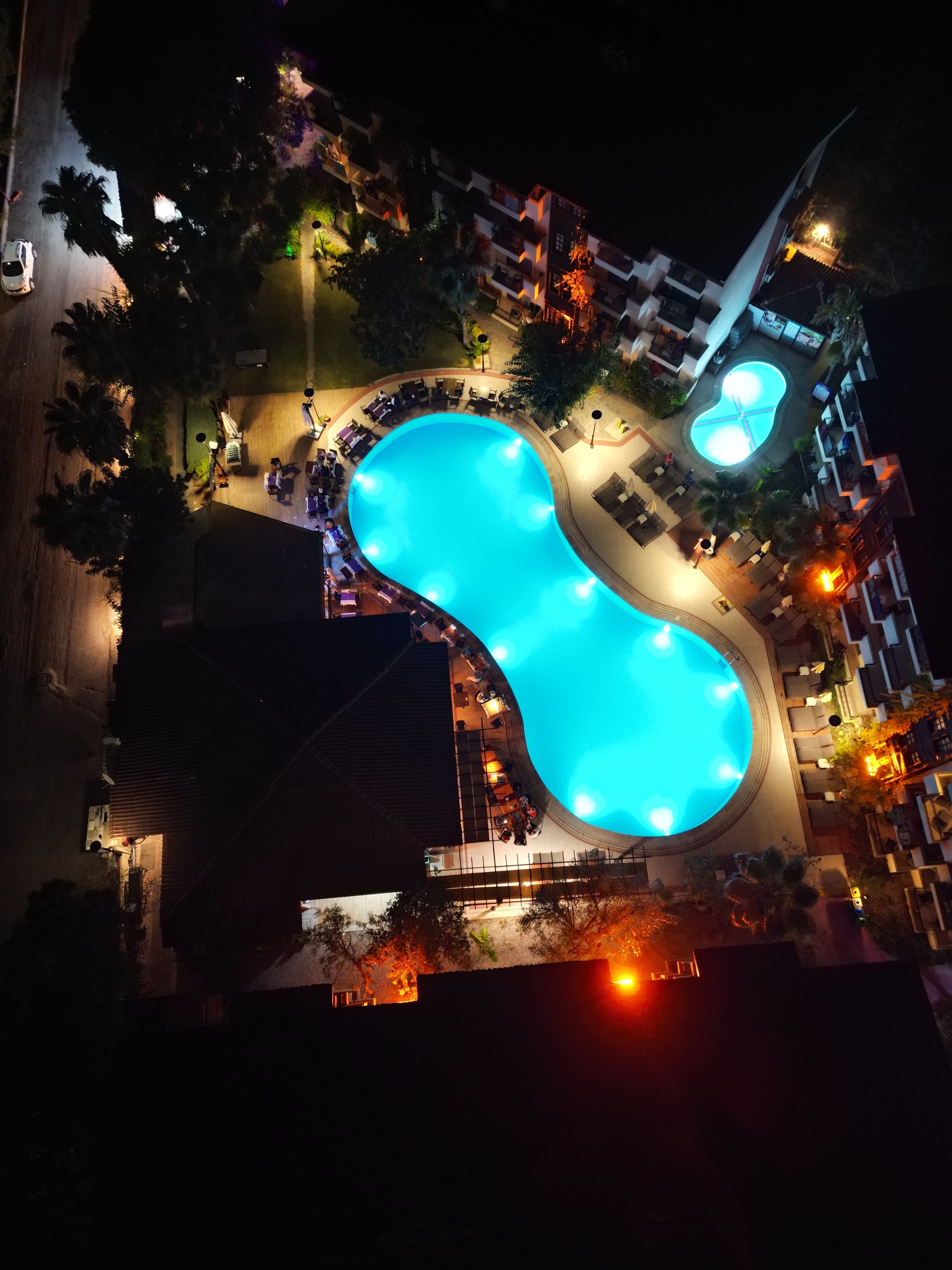Luchtbeeld van een hotel met een zwembad in een bijzondere vorm, omringd door terrasjes en verlicht met sfeerverlichting.