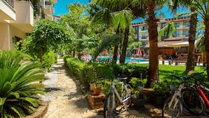 A pathway lined with potted plants and bicycles, leading to a pool area with umbrellas and a building in the background. - Lemas Suite Hotel  (Manavgat)