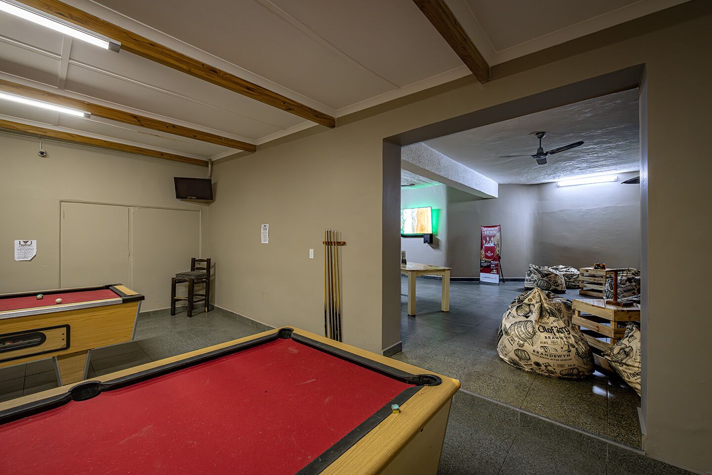 A pool table with a red felt surface, a wooden pool table, and a ceiling fan in a room with beige walls and a wooden ceiling beam.
