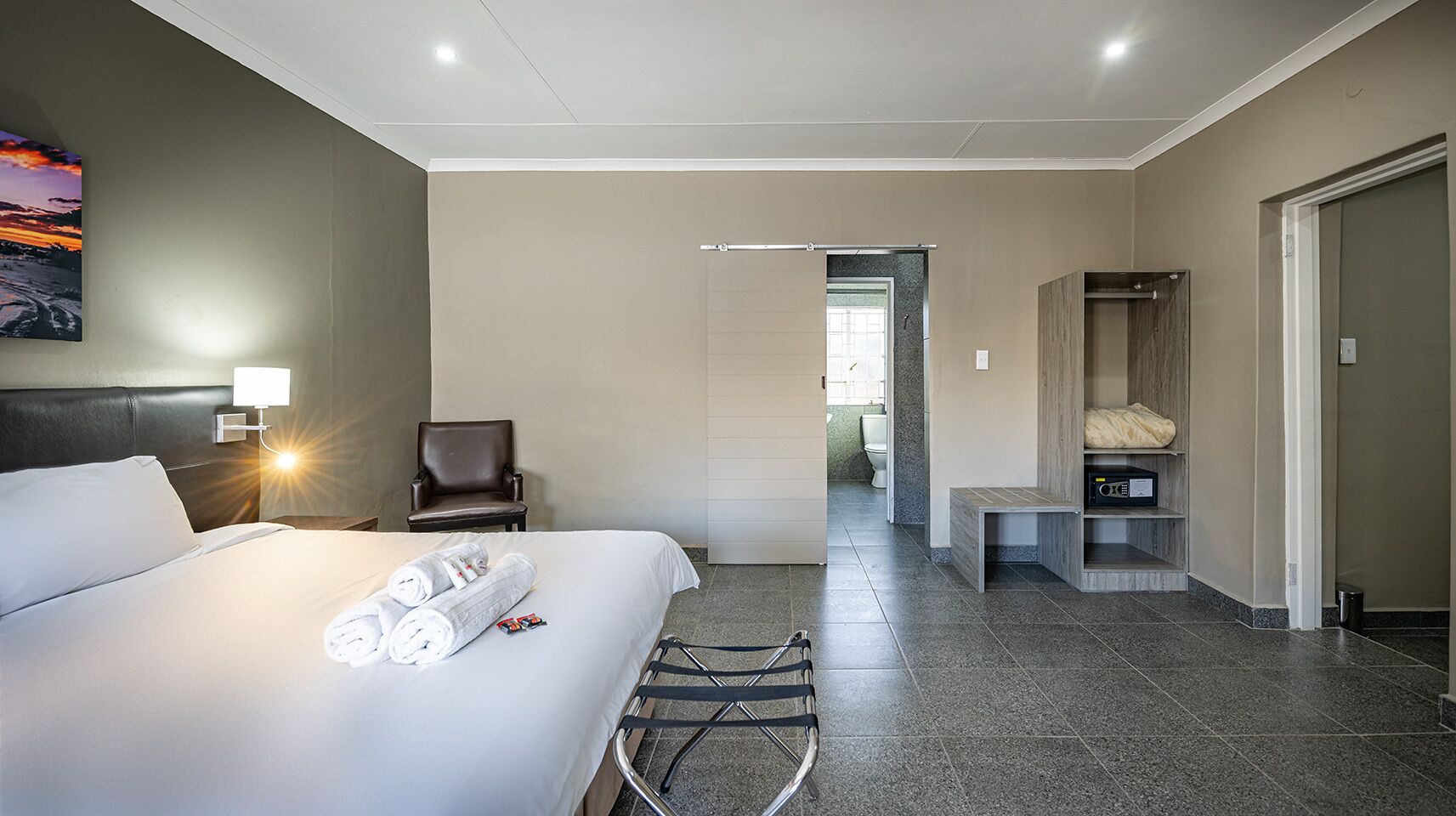 Une chambre d’hôtel avec un lit, une chaise, un bureau avec une radio, un miroir et une vue sur la salle de bain.