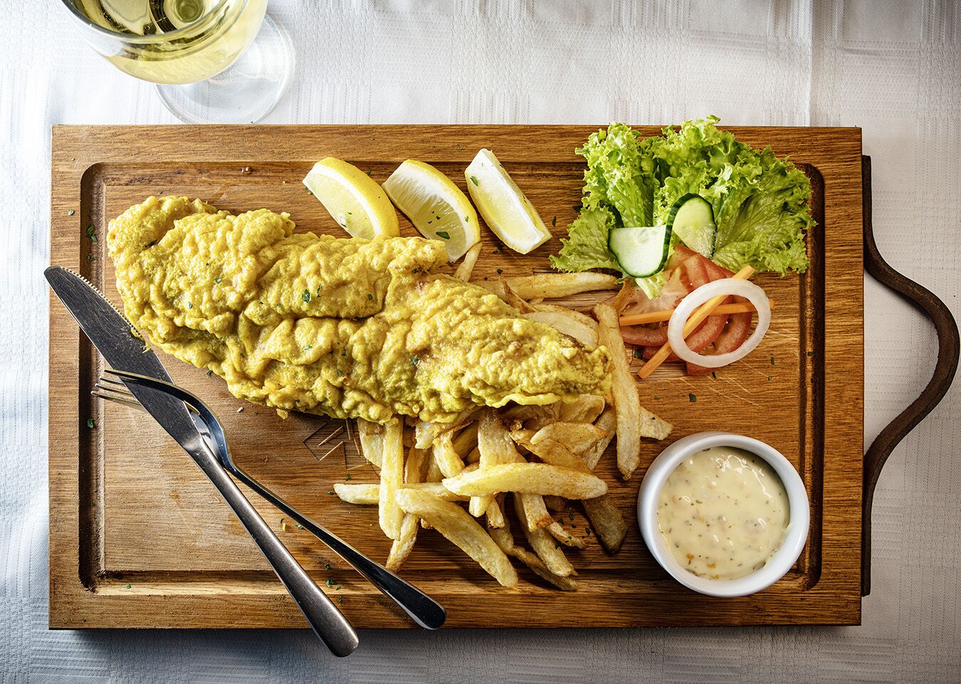 Un plato de pescado y papas fritas con cuñas de limón, lechuga, pepino, tomate y cebolla, junto con un recipiente pequeño de salsa sobre una tabla de madera.