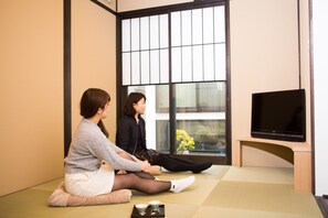 Dos personas sentadas sobre esteras de tatami en una habitación con una televisión y una ventana que ofrece vistas a un edificio y un árbol.