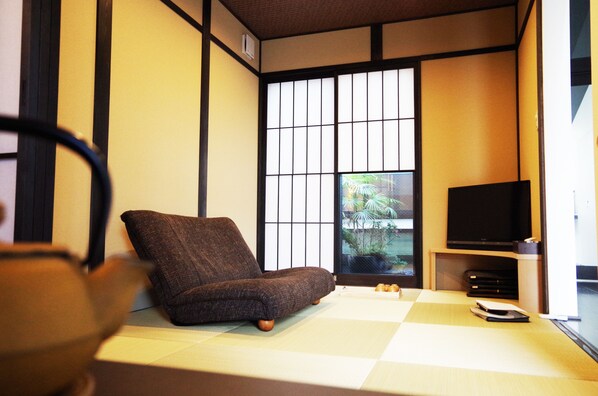 Una habitación japonesa tradicional con suelo de tatami, un sofá marrón bajo, un televisor sobre un soporte de madera y un panel corredizo shoji.