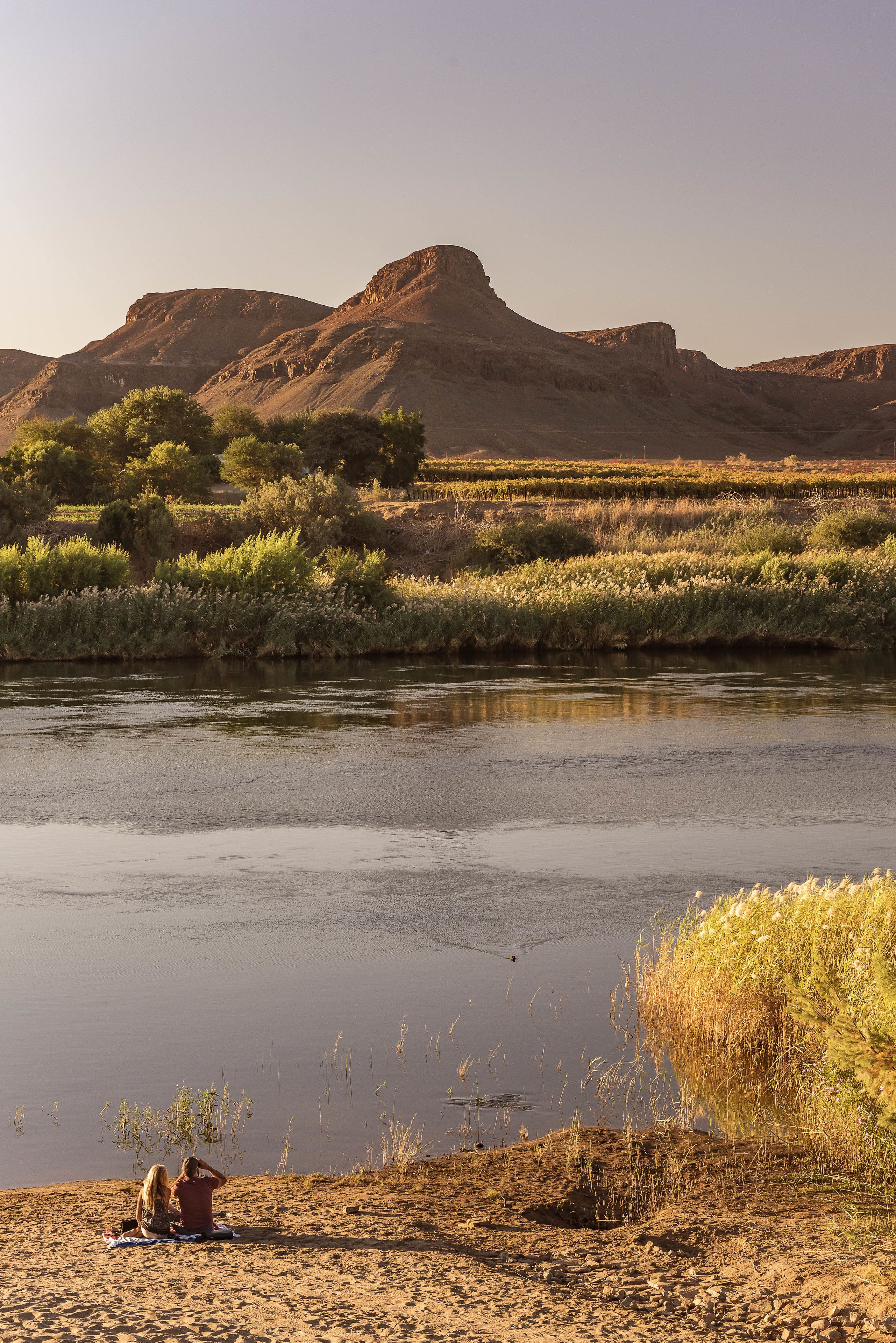 A river with a sandy bank and two people sitting on a blanket.