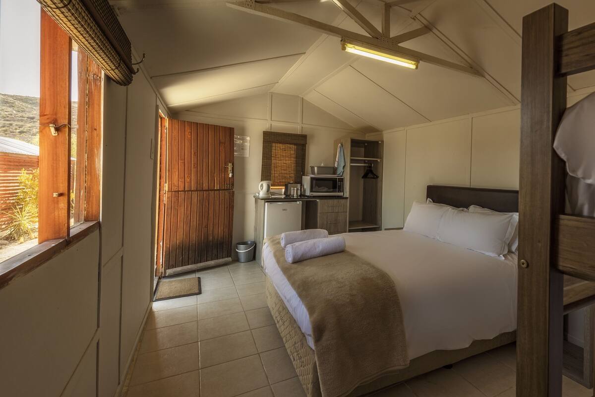 A bedroom with a large bed, a wooden headboard, a small kitchenette, and a view of the outdoors.