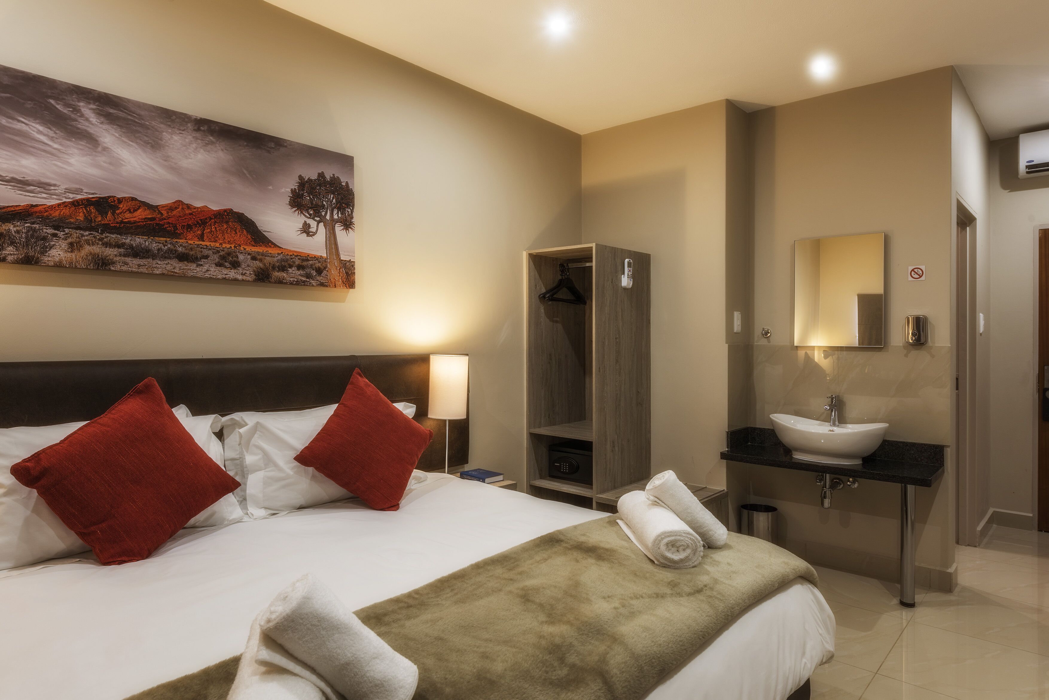 A hotel room with a bed, red pillows, a painting, a wardrobe, a sink, and a mirror.