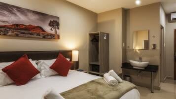 A hotel room with a bed, red pillows, a painting, a wardrobe, a sink, and a mirror.