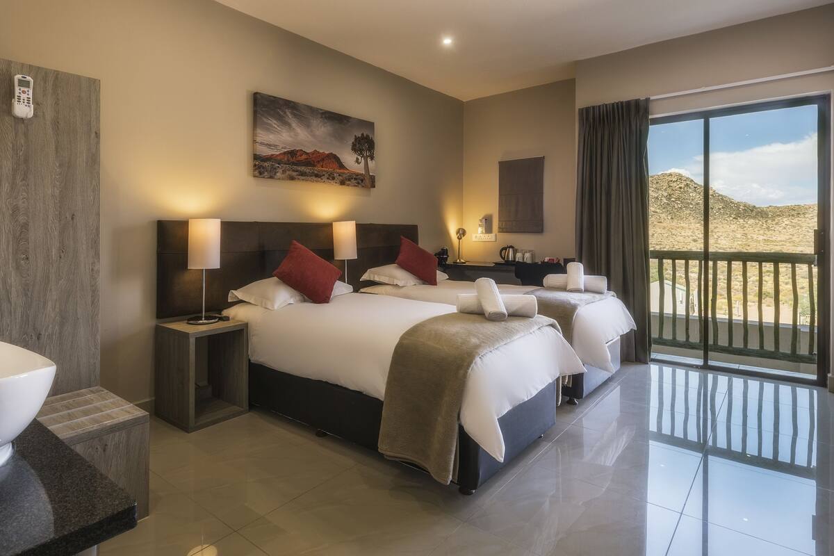 A hotel room with a large white sink, two beds with red pillows, a painting on the wall, and a balcony with a view of the desert.