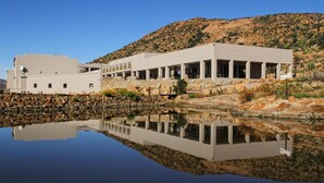 A modern building with a reflective pool in front, surrounded by rocky terrain and a clear sky. - Springbok Inn by Country Hotels (Nama Khoi)