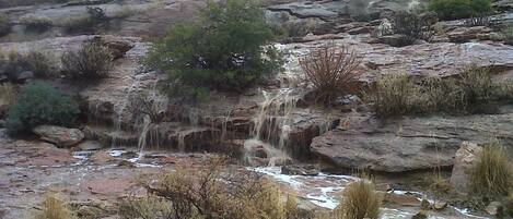 A rocky landscape with a waterfall, sparse vegetation, and a forested hill in the background.