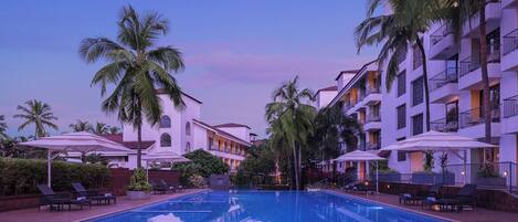 A swimming pool with lounge chairs and umbrellas, surrounded by palm trees and a multi-story building with balconies.