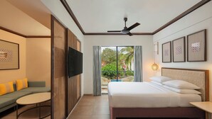 Habitación de hotel con cama, sofá, televisor, balcón con vistas a zonas verdes y ventilador de techo.