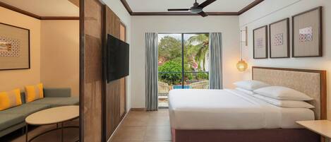 A hotel room with a bed, a sofa, a TV, a balcony with a view of greenery, and a ceiling fan.