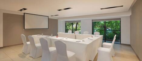 A conference room with a long table, white chairs, a projector screen, and large windows with a view of greenery.