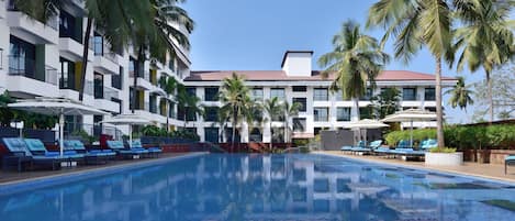 A swimming pool with lounge chairs and umbrellas, surrounded by palm trees and a multi-story building with balconies.