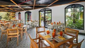 Un restaurante con mesas y sillas de madera, piso de baldosas y ventanas amplias con vistas a zonas verdes.