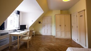 A room with a sloped ceiling, a desk with a computer, a chair, and a bed. There are two doors and a window with curtains. - Tyn Yard Residence (Praha)
