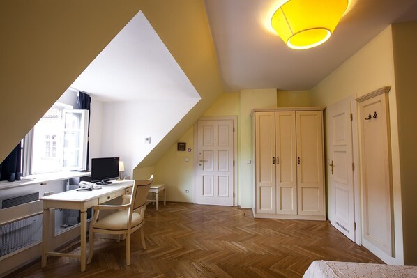 A room with a sloped ceiling, a desk with a computer, a chair, and a bed. There are two doors and a window with curtains. - Tyn Yard Residence (Praha)