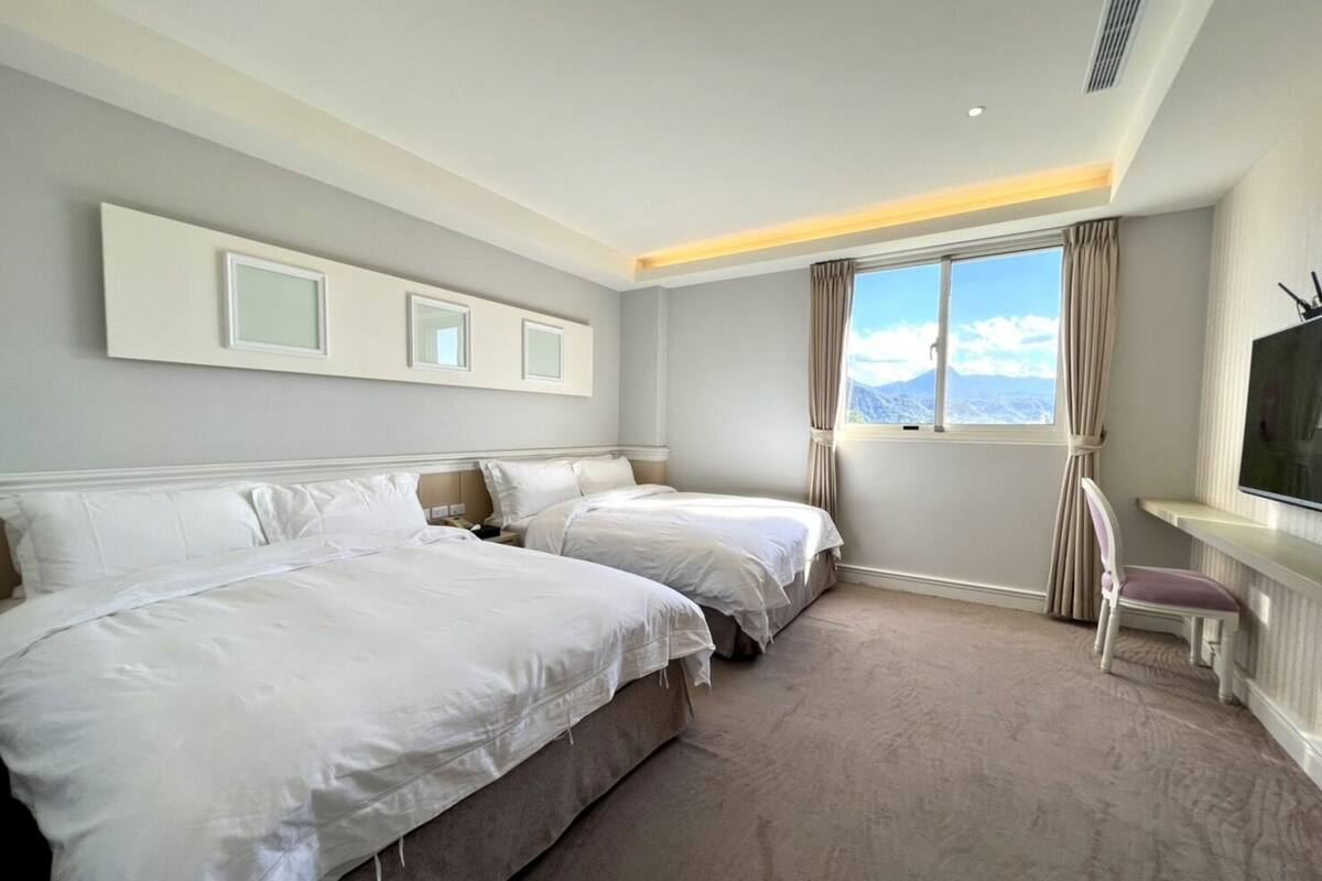 A hotel room with two beds, a TV, a window with a mountain view, and a chair.