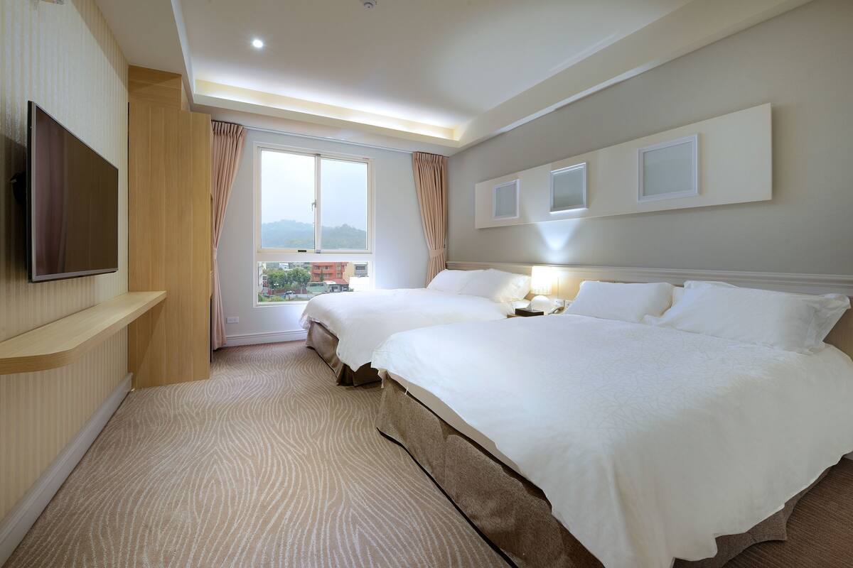 A hotel room with two beds, a TV, a large window with curtains, and a view of the outside.
