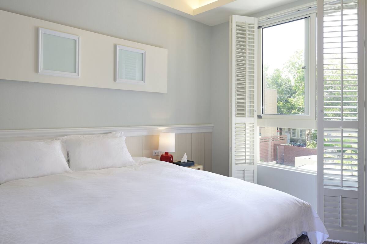 A neatly made bed with white linens in a room with a large window and white shutters.