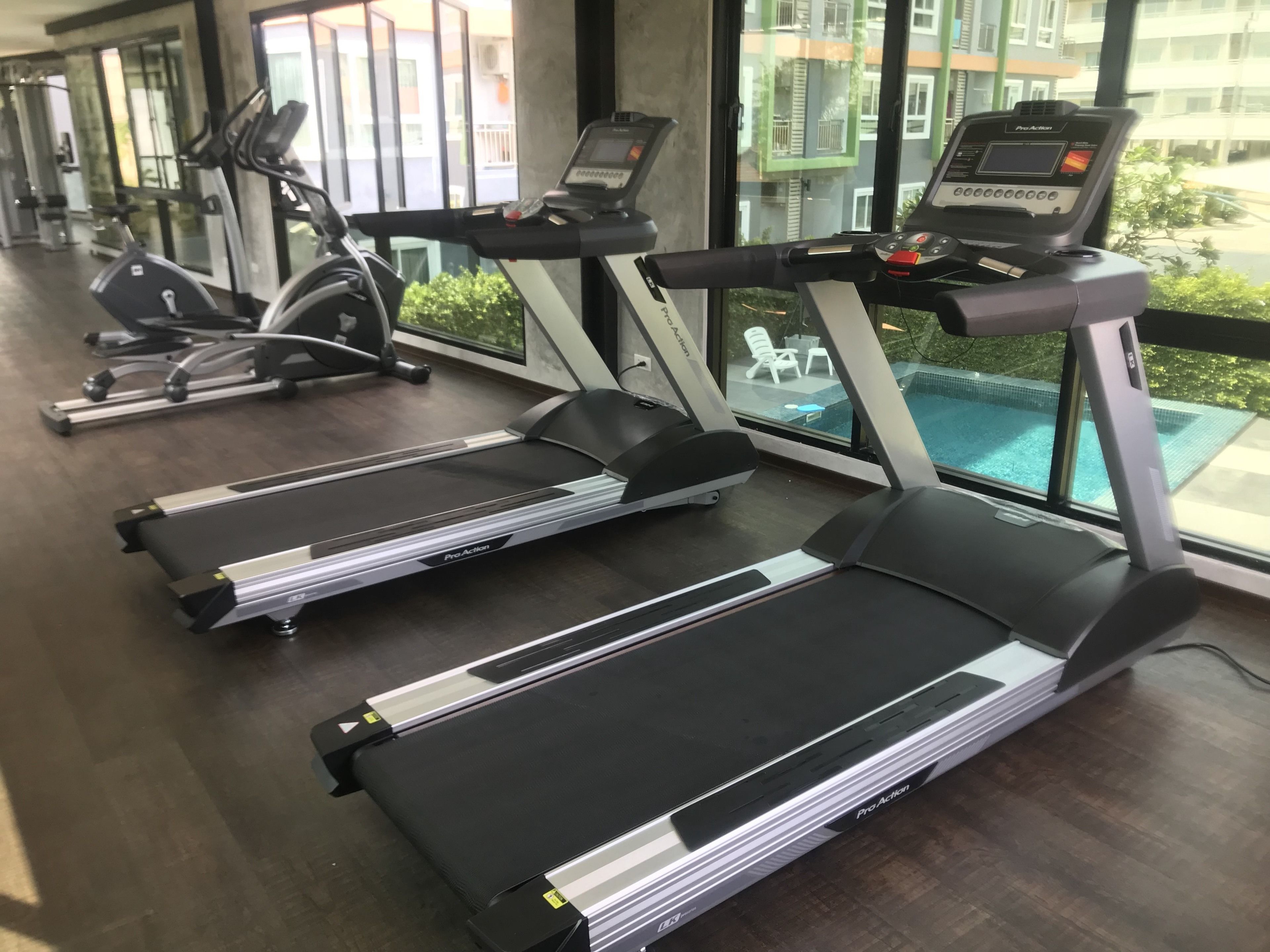 A row of Pro Action treadmills in a gym with large windows overlooking a pool and residential buildings.