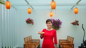 A person in a red dress holding a plate with food, standing in a room with wooden chairs, hanging orange lanterns, and potted plants. - Minh Phat Homestay (Da Nang)