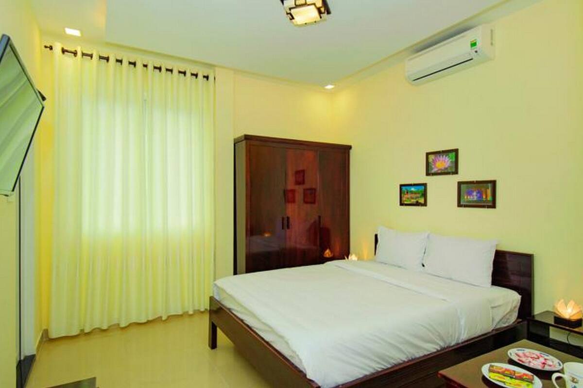 A hotel room with a bed, a TV, a wardrobe, an air conditioner, and a nightstand with a lamp and a flower.
