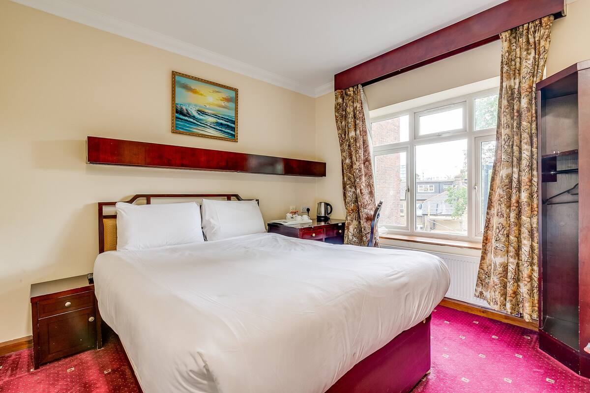 A hotel room with a large bed, a nightstand, a painting on the wall, and a window with curtains.