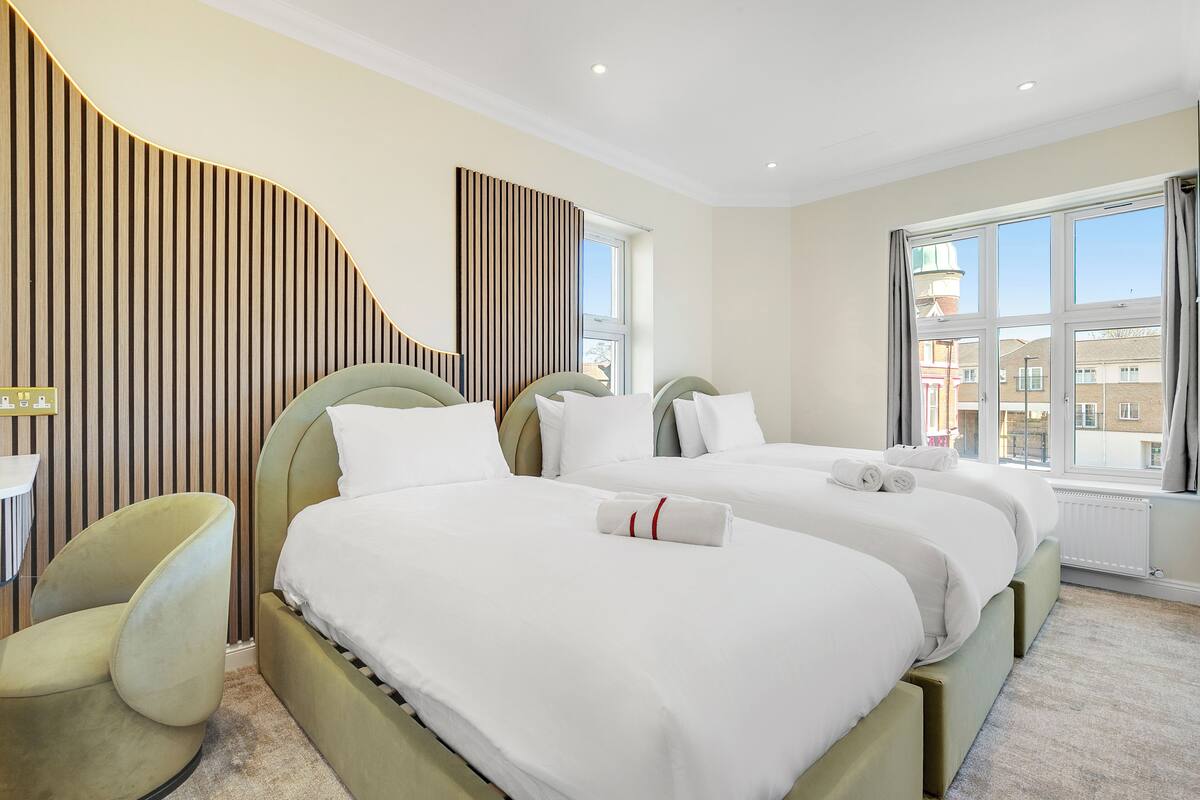 A hotel room with two beds, a green armchair, a window with a view of buildings, and a wooden panel wall.