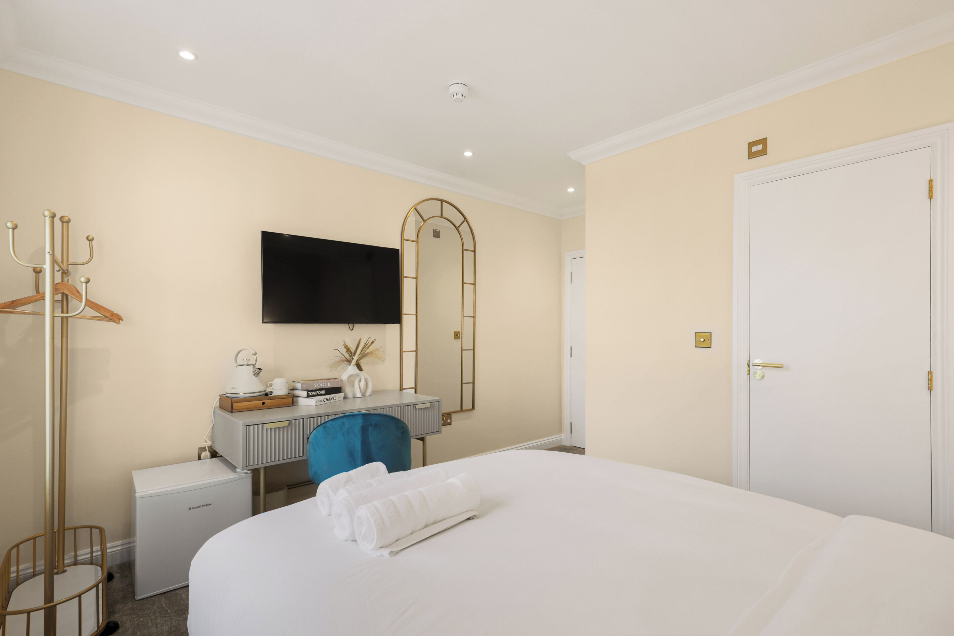 Quarto de hotel com cama, escrivaninha com cadeira, televisão, armário e cabideiro.
