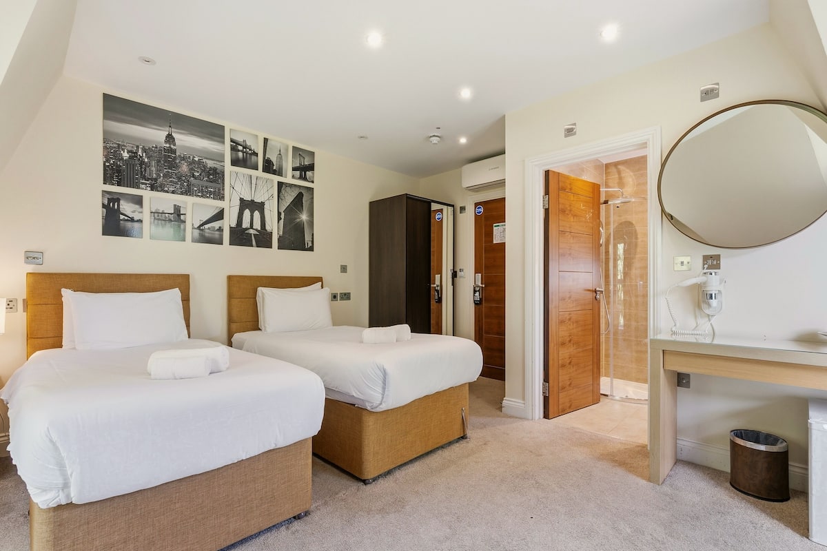A hotel room with two beds, a wooden wardrobe, a bathroom with a mirror, and a cityscape wall art.