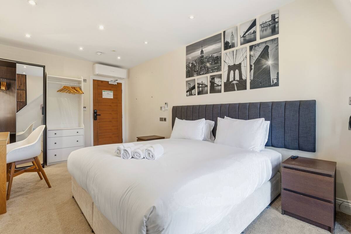 A hotel room with a large bed, a desk, a chair, a wardrobe, and a wall with black and white cityscape photos.