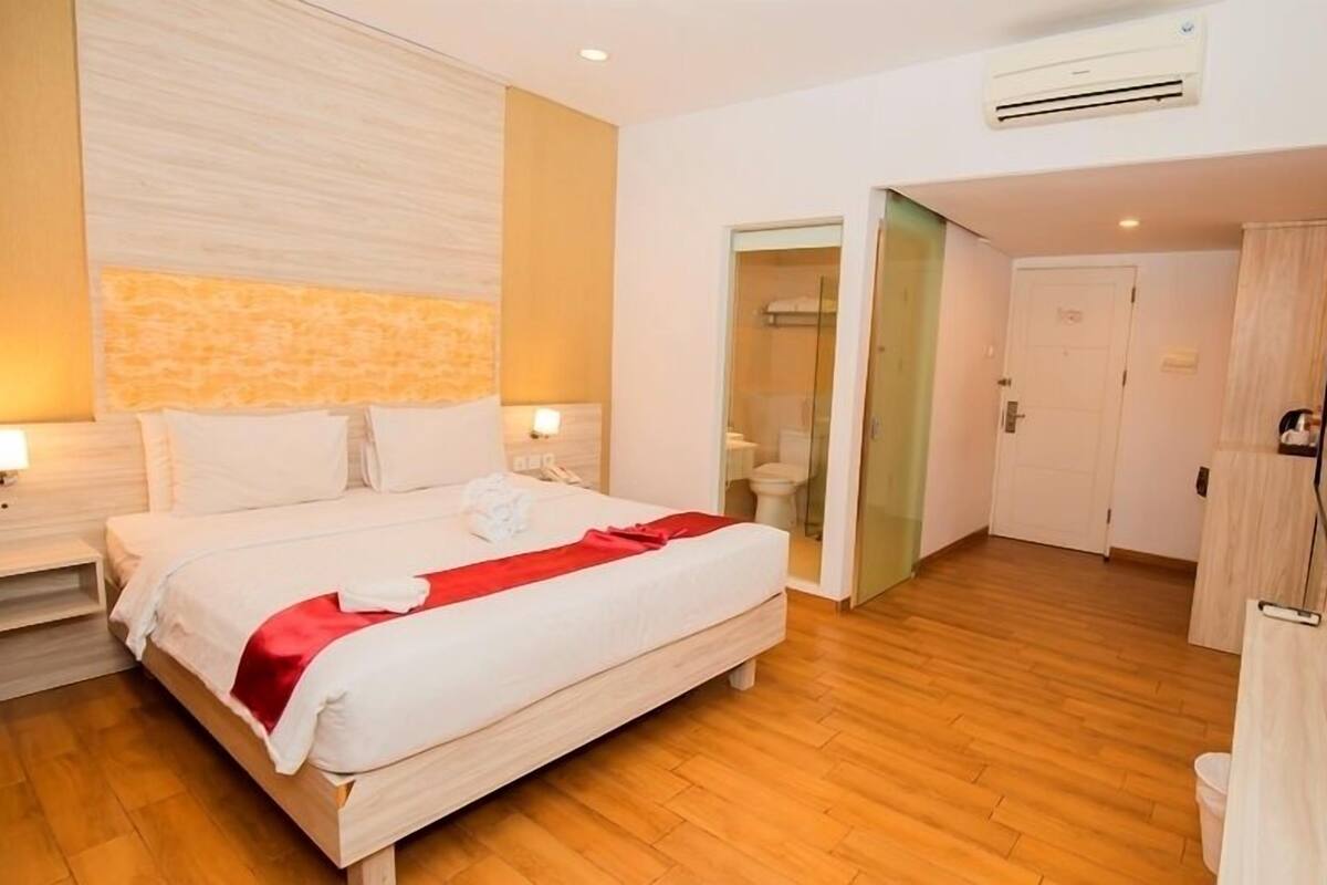 Een moderne hotelkamer met een groot bed, houten vloeren, een ingebouwde televisie en een aangrenzende badkamer, zichtbaar door een geopende deur.