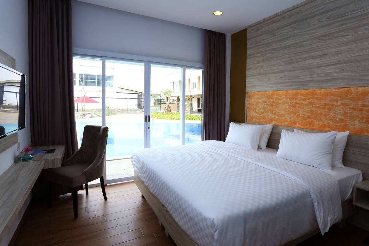 Hotelkamer met een groot bed, een bureau met stoel en uitzicht op een zwembad door een glazen deur.