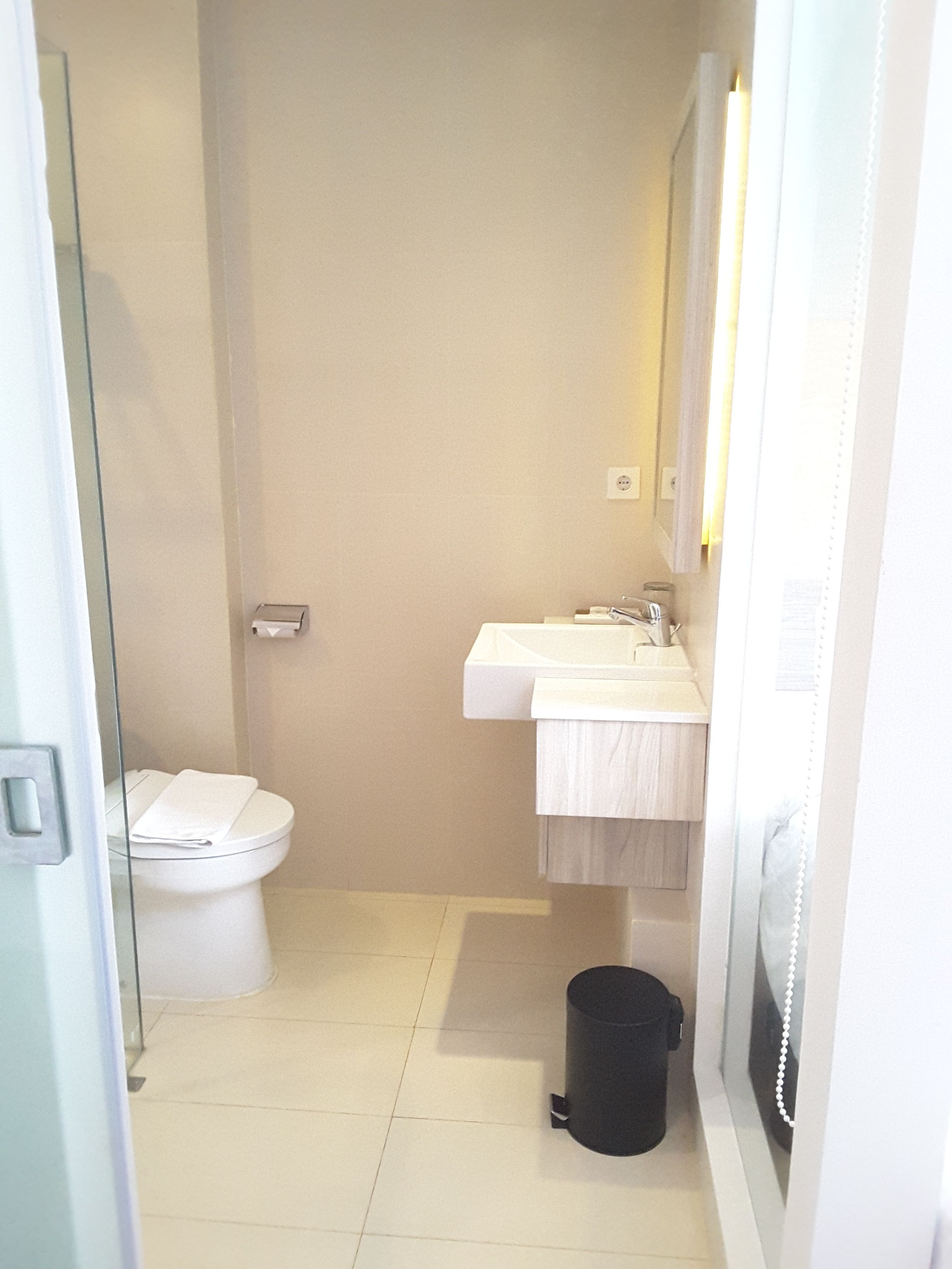 A small, narrow bathroom with a toilet, sink, and a trash bin.