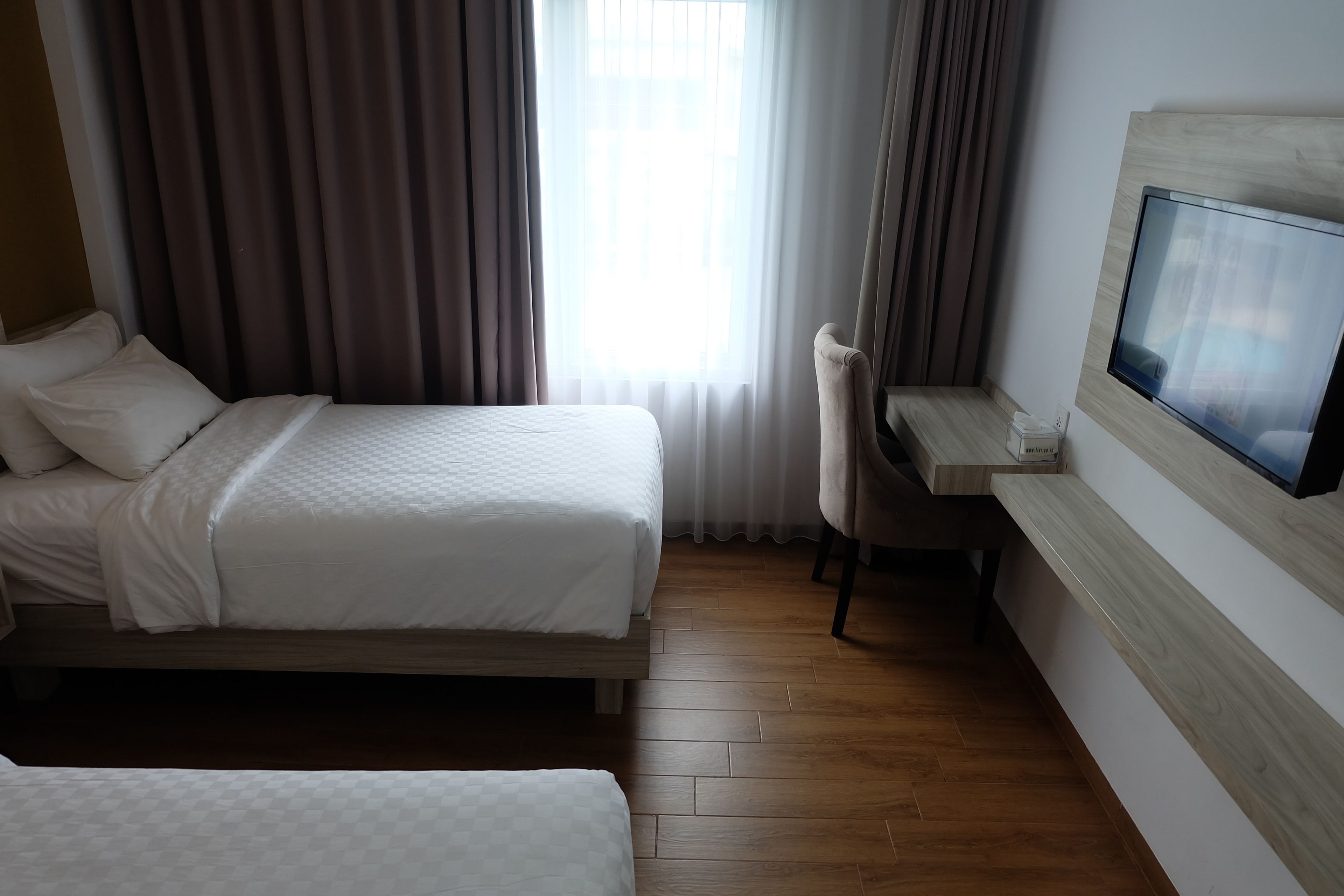 A hotel room with two beds, a desk with a chair, a TV, and a large window with curtains.