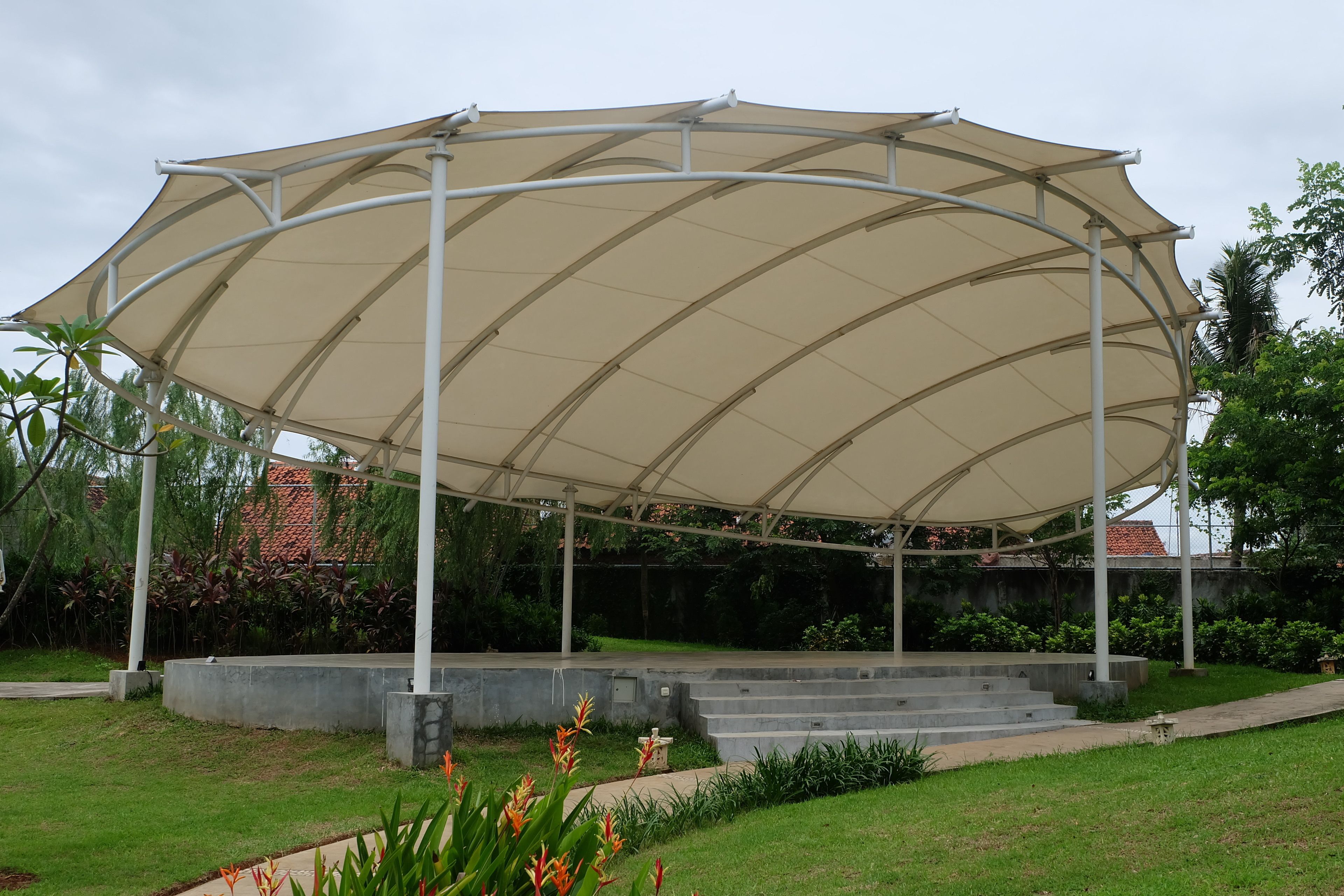 A covered pavilion with a curved roof structure, surrounded by a landscaped garden with steps leading up to it.